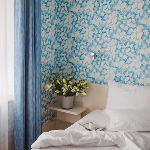 Blick auf ein Hotelbett neben einem bunten Blumenstrauß und vor weiß-türkis geblümter Tapete